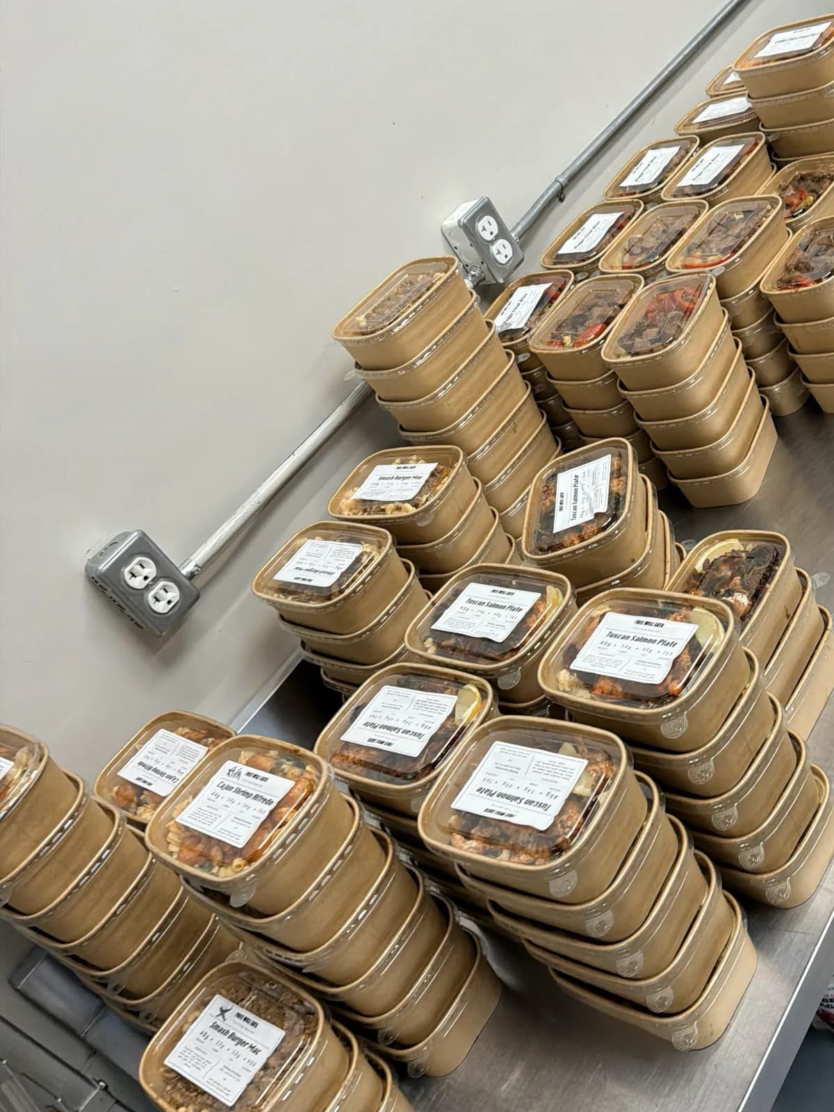 Dozens of kraft boxes stacked down the steel counter, each sealed and labelled with its meal name and macros