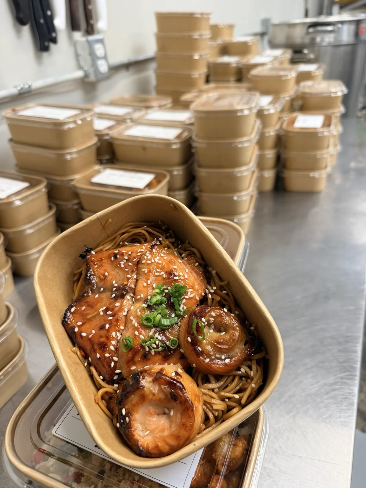 A kraft box of glazed salmon on noodles held over the kitchen's steel counter, with labelled boxes stacked down the line behind it